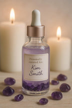 May include: A clear glass bottle with a white dropper and a rose gold cap, filled with a shimmering purple liquid and amethyst stones. The label reads "Personally curated for: Kim Smith." Two lit candles and amethyst stones surround the bottle.